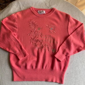 May include: A coral pink long-sleeve sweater featuring a crew neckline and ribbed cuffs and hem. The front displays an embroidered floral design with leaves and a single small circular embellishment. A white brand label is sewn inside the collar.