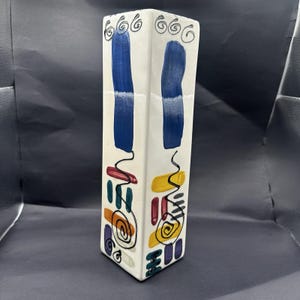 May include: A tall, rectangular ceramic vase with a white base. The surface features two vertical blue brushstrokes, black hand-drawn spiral and squiggle line art, and a series of small, colorful horizontal paint dabs in yellow, red, and teal.