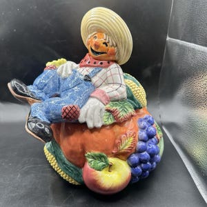 Peut inclure: Un pot à biscuits en céramique en forme d'épouvantail assis sur une récolte composée d'une grosse citrouille orange, de maïs jaune, de pommes rouges et jaunes et de raisins violets. L'épouvantail porte un chapeau de paille, une chemise à carreaux.