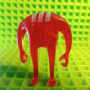 May include: A translucent red figurine with a minimalist design. The figure has a rectangular head with three white stripes, two arms, and two legs. The background is a textured green surface.