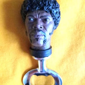 May include: A bottle opener featuring a sculpted head with dark skin and a black afro. The head is attached to a black rubber stopper and a silver bottle opener. The background is a solid yellow.