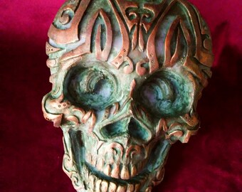 May include: A green and gold skull sculpture with intricate details and a decorative design. The skull is made of a textured material and has a distressed finish.