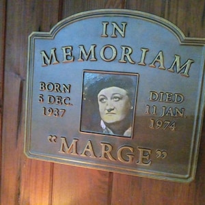 May include: A brass plaque with gold lettering reads "In Memoriam" with a photo of a woman and the text "Born 5 Dec. 1937 Died 11 Jan. 1974 "Marge""
