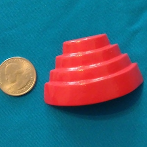 May include: A red plastic toy shaped like a tiered cake. The toy is about the size of a US quarter.