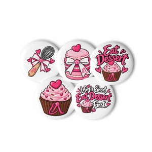 May include: A set of five round white buttons featuring pink-themed dessert illustrations. Designs include a whisk with a bow, a stacked macaron, a cupcake, and text reading Eat Dessert and Life Is Short Eat Dessert First.