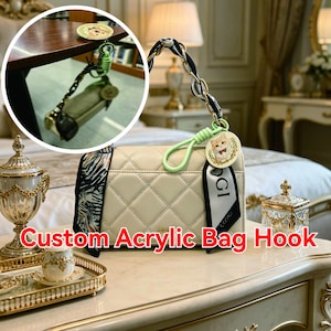 May include: Cream-colored quilted handbag with a black and white patterned scarf and a decorative chain. A custom acrylic bag hook with a cat illustration is attached. The text "Custom Acrylic Bag Hook" is displayed.