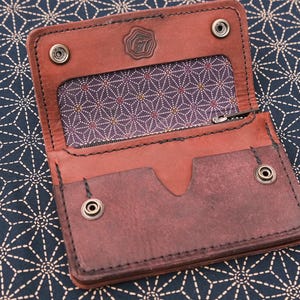 May include: Open, brown leather wallet with a patterned interior and snap closures. The wallet has a small logo on the top flap and is displayed on a dark blue surface with a white geometric pattern.