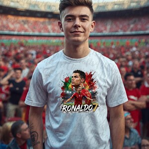 May include: A white short-sleeved t-shirt featuring a graphic print of soccer player Cristiano Ronaldo in a red jersey, set against a colorful abstract background with red and green paint splatters and the text RONALDO 7 in bold white lettering.