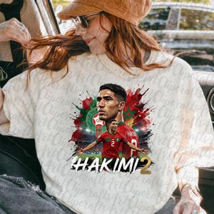 May include: A white t-shirt featuring a graphic print of a soccer player in a red jersey with the number 2. The design includes red and green watercolor splashes, a soccer emblem, and the text HAKIMI 2 in bold black and gold lettering.