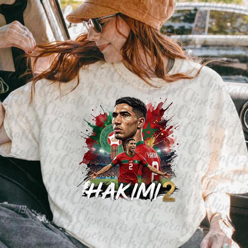 May include: A white t-shirt featuring a graphic print of a soccer player in a red jersey with the number 2. The design includes red and green watercolor splashes, a soccer emblem, and the text HAKIMI 2 in bold black and gold lettering.