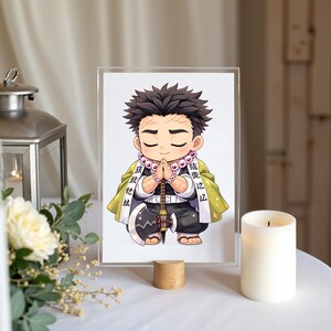 May include: A framed illustration of a chibi-style anime character in a prayer pose. The character has dark hair, a green and yellow cloak, and a beaded necklace. The image is displayed on a wooden stand with a white candle and a lantern in the background.