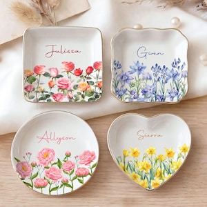 Puede incluir: Cuatro platos decorativos para anillos de diferentes formas: cuadrado, festoneado, redondo y en forma de corazón. Cada plato presenta un diseño floral con los nombres: Julissa, Gwen, Allyson y Sierra. Los platos tienen bordes dorados y están sobre una superficie blanca.