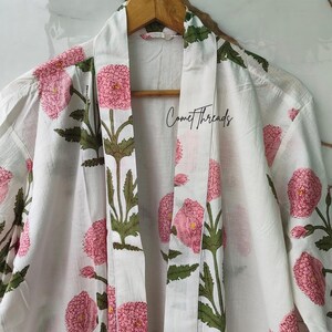 May include: White cotton robe featuring a floral design of pink flowers and green leaves. The robe is suspended from a wooden hanger. The text "Comet Threads" is printed on the robe.