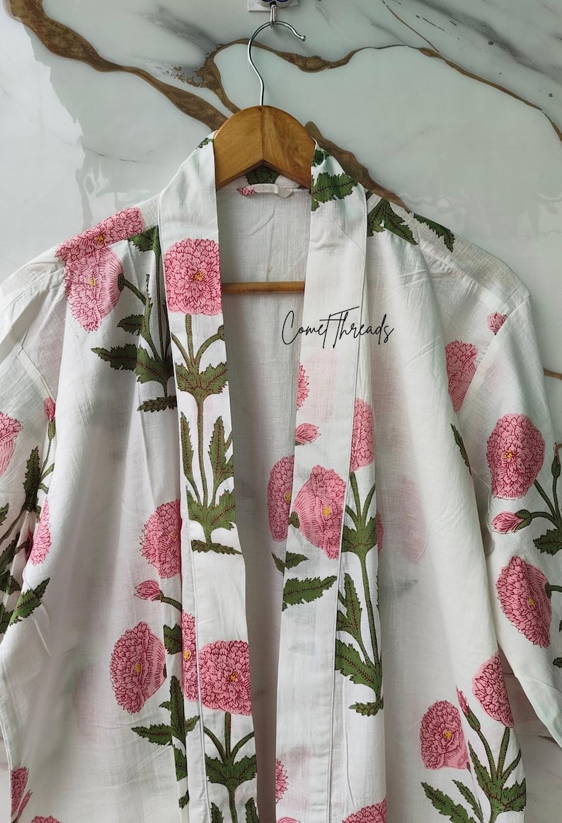 May include: White cotton robe featuring a floral design of pink flowers and green leaves. The robe is suspended from a wooden hanger. The text "Comet Threads" is printed on the robe.