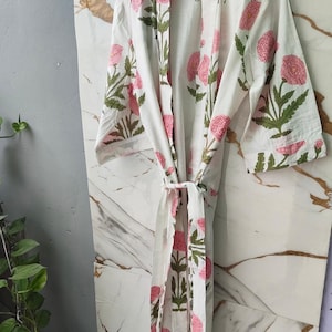 May include: White cotton robe featuring a pink and green floral design. The robe has a tie closure and is suspended from a wooden hanger. The text "Comet Threads" is visible on the robe. The robe is set against a marble-effect backdrop.