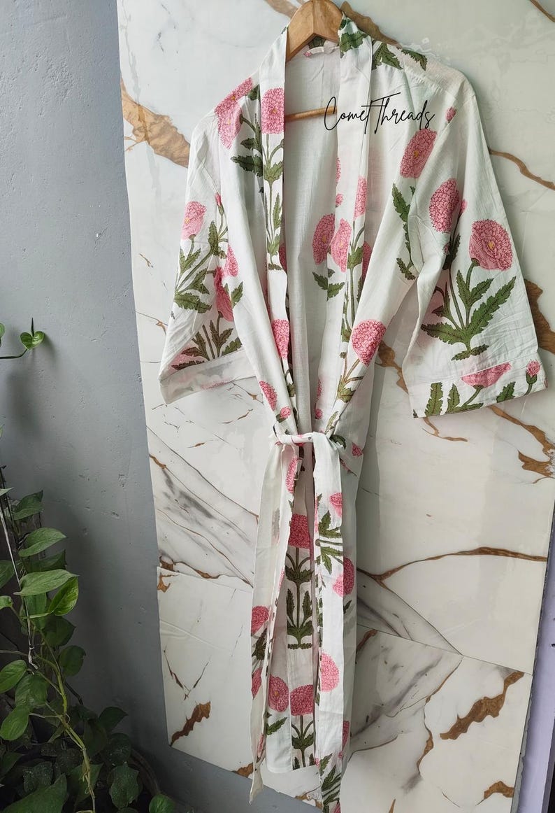 May include: White cotton robe featuring a pink and green floral design. The robe has a tie closure and is suspended from a wooden hanger. The text "Comet Threads" is visible on the robe. The robe is set against a marble-effect backdrop.