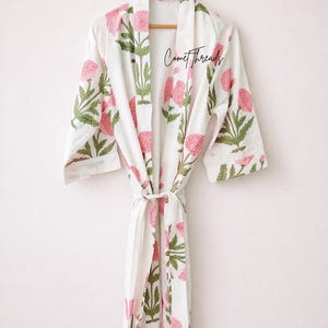 May include: White robe with a pink floral pattern and green leaves, hanging on a wooden hanger. The robe has a tie closure and the text "Comet Threads" is visible on the front.