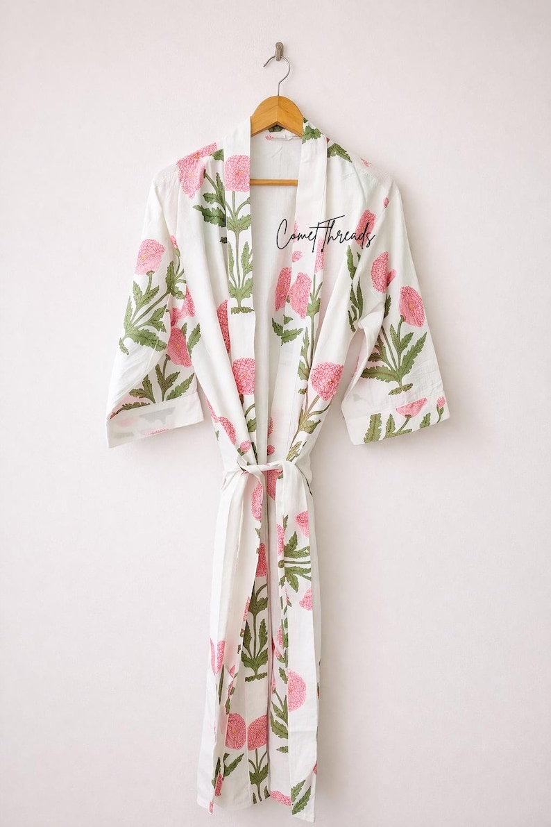 May include: White robe with a pink floral pattern and green leaves, hanging on a wooden hanger. The robe has a tie closure and the text "Comet Threads" is visible on the front.