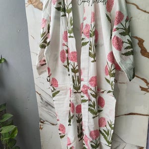 May include: White robe with a floral pattern of pink flowers and green leaves. The robe has long sleeves and pockets. The text "CometThreads" is visible. The robe is hanging on a wooden hanger.