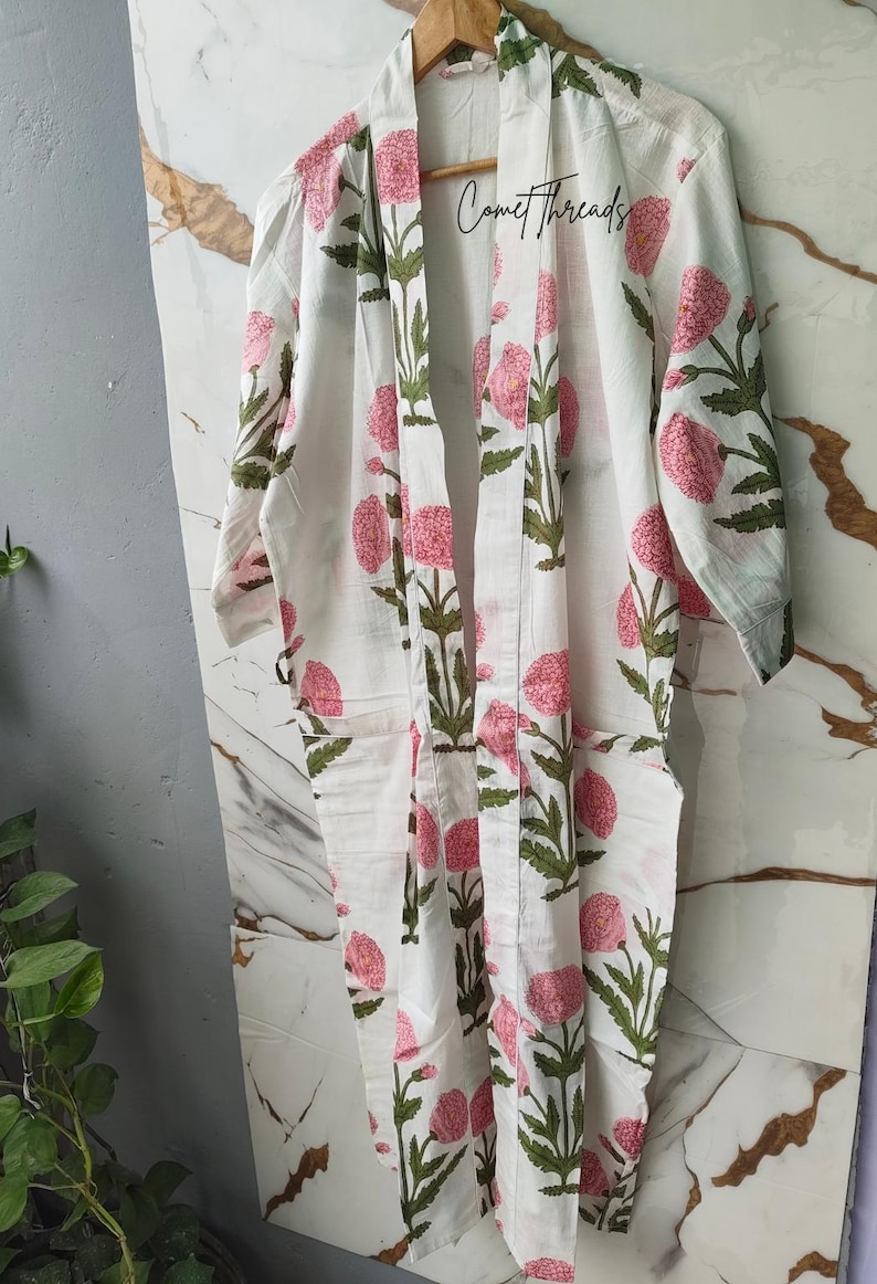 May include: White robe with a floral pattern of pink flowers and green leaves. The robe has long sleeves and pockets. The text "CometThreads" is visible. The robe is hanging on a wooden hanger.