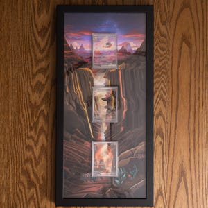 May include: A rectangular black frame containing three vertically aligned trading cards displayed over a printed background of a volcanic canyon landscape with a sunset sky. The cards feature illustrations of fire-type creatures in a rocky terrain.