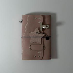 May include: A dusty rose leather journal cover featuring a silver metal binder clip, a star-shaped suede charm, a black elastic band, and a silver paperclip. The cover is secured with silver rivets and includes a small black heart-shaped charm.