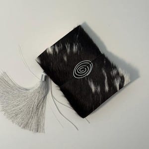 以下が含まれることがあります： 白と黒の牛革で覆われた長方形のジャーナル。表紙の中央には、細い黒いゴムバンドに固定された銀色の金属製スパイラルエンブレムがあしらわれています。側面には長いライトグレーのタッセルが付いています。
