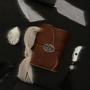 Könnte beinhalten: Ein braunes, strukturiertes Notizbuch mit einem spiralförmigen Metallverschluss, umwickelt mit einem hellbeigen Band. Das Notizbuch ist umgeben von einem silbernen muschelförmigen Objekt, einem schwarzen Kamm und einem silbernen Werkzeug.