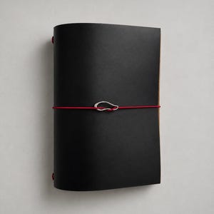 May include: A black leather journal cover featuring a vibrant red elastic band wrapped horizontally around the center. A small, irregular silver-toned metal charm is threaded onto the red elastic, securing the cover closed against the pages.