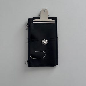 Może przedstawiać: Czarny skórzany notatnik z srebrnym zapięciem na klips. Notatnik jest zabezpieczony czarną gumką i srebrnym sercem. Srebrne zawieszki w kształcie gwiazd wiszą z boku. Dołączony jest srebrny spinacz do papieru.