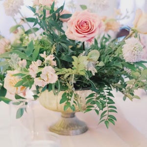 Puede incluir: Arreglo floral con una gran rosa rosa como punto focal, rodeada de otras flores rosas y blancas y follaje verde. El arreglo está en un jarrón con pedestal dorado, creando una exhibición elegante.