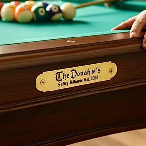 May include: A close-up of a pool table with a green felt surface and wooden frame. Several billiard balls are visible, along with a cue stick and a brass plaque that reads "The Donahue's Salty Billiards Est. 2026".