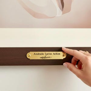May include: Abstract artwork in a neutral colour scheme, framed with a dark brown wooden frame. A gold-coloured nameplate on the frame reads "Andrada Larisa Achim" with a signature. The artwork features beige and brown shapes. Displayed on a white wall.