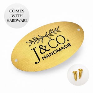 Peut inclure: Une étiquette ovale en métal doré avec le texte "J & CO. HANDMADE" en noir. Un motif décoratif de branche feuillue est au-dessus du texte. L'étiquette comprend deux trous pré-percés et est livrée avec deux vis dorées. Le texte "COMES WITH HARDWARE" est également présent.