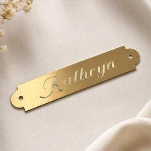 May include: A rectangular gold-toned metal nameplate with rounded ends and a small circular hole on each side. The name Kathryn is engraved in a cursive script font across the center of the plate, featuring a smooth, polished finish.