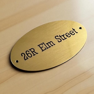 Puede incluir: Placa de dirección ovalada de latón con el texto "26R Elm Street" en una fuente oscura con serifas. La placa tiene dos pequeños agujeros, uno en cada extremo. El fondo es una veta de madera clara.
