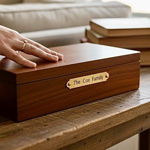 May include: A rectangular wooden box with a dark brown finish. A gold-colored plaque on the front reads "The Cox Family." The box is on a wooden surface, with a hand resting on top. Books are in the background.