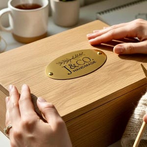 Może przedstawiać: Jasna drewniana skrzynka ze złotą owalną plakietką z napisem "J & CO. HANDMADE" czarnymi literami. Skrzynka jest prostokątna i wydaje się zamknięta. Obraz przedstawia zbliżenie skrzynki z dłońmi spoczywającymi na niej.