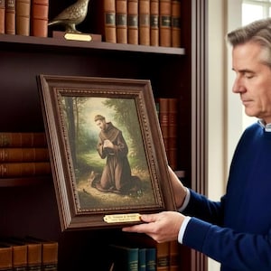 May include: A framed painting of a man in a brown robe kneeling in a forest setting. The painting has a dark brown wooden frame. The painting is being held in front of a bookshelf filled with books.