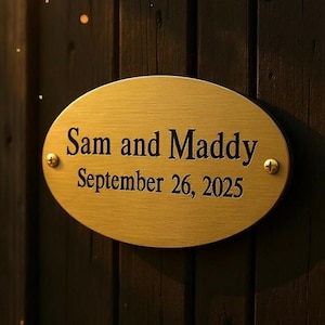 Puede incluir: Placa ovalada dorada con el texto grabado "Sam and Maddy September 26, 2025." La placa está montada sobre una superficie de madera oscura con dos tornillos pequeños. El texto está en una fuente serif clásica.