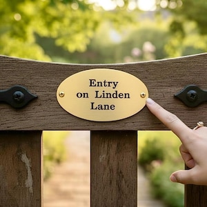 May include: A wooden bench with a gold oval plaque that reads "Entry on Linden Lane." The plaque is affixed to the bench with small gold screws. The bench is made of dark brown wood and has decorative black metal hardware.