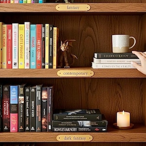 Puede incluir: Una estantería de madera con libros, una figura de dragón y una vela. Las estanterías están etiquetadas como "fantasy", "contemporary" y "dark fantasy". Una mano alcanza una taza moteada sobre una pila de libros.