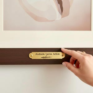 Puede incluir: Obra de arte enmarcada con un diseño suave y abstracto en tonos neutros. La obra está montada con un passe-partout blanco y un marco marrón oscuro. Una placa dorada en el marco dice "Andrada Larisa Achim" con una firma.