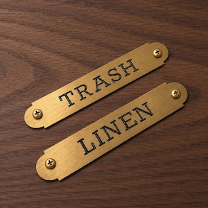Könnte beinhalten: Zwei rechteckige Messingschilder mit den Wörtern "TRASH" und "LINEN" in schwarzer Schrift. Jedes Schild ist mit zwei Messingschrauben befestigt. Der Hintergrund ist eine dunkle Holzmaserung.