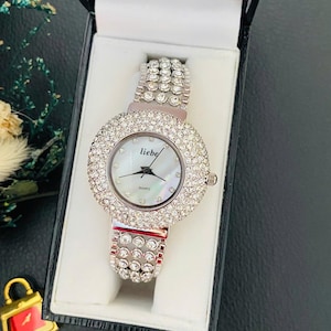 May include: A silver wristwatch with a round, crystal-encrusted face and matching band. The watch face has a mother-of-pearl effect and the word "liebe" is printed on the face. The watch is presented in a black box.