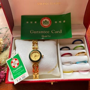 May include: A gold-tone Charmex analogue quartz watch with a dark green bezel and a decorative link bracelet, presented in a red box with a guarantee card and a set of ten colourful interchangeable rings stored in a white velvet insert.