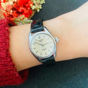 May include: A vintage Universal Geneve Polerouter automatic wristwatch with a silver case and a black leather strap. The watch has a cream-coloured face with silver hands and markers. The watch is displayed on a wrist.