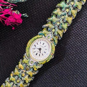 Puede incluir: Un reloj vintage con una pulsera articulada. La esfera es redonda, crema con agujas y marcadores negros. La pulsera y el bisel presentan un patrón verde y amarillo. Un accesorio de moda.