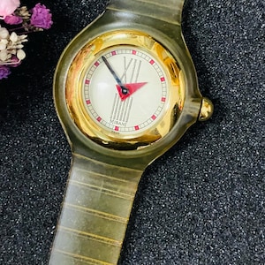 Puede incluir: Un reloj de pulsera con correa verde oliva translúcida y bisel circular dorado. La esfera color crema presenta una manecilla de hora triangular roja, un minutero negro, el texto MISANI y el número romano XII en la parte superior.