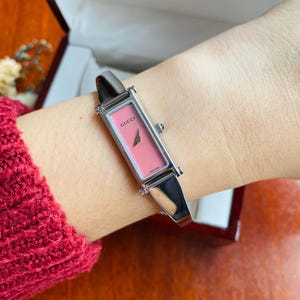 May include: A Gucci wristwatch with a rectangular silver-tone metal case and a solid pink dial. The watch features a polished silver-tone bangle-style bracelet, a small side crown, and the Gucci logo printed in black on the face of the dial.